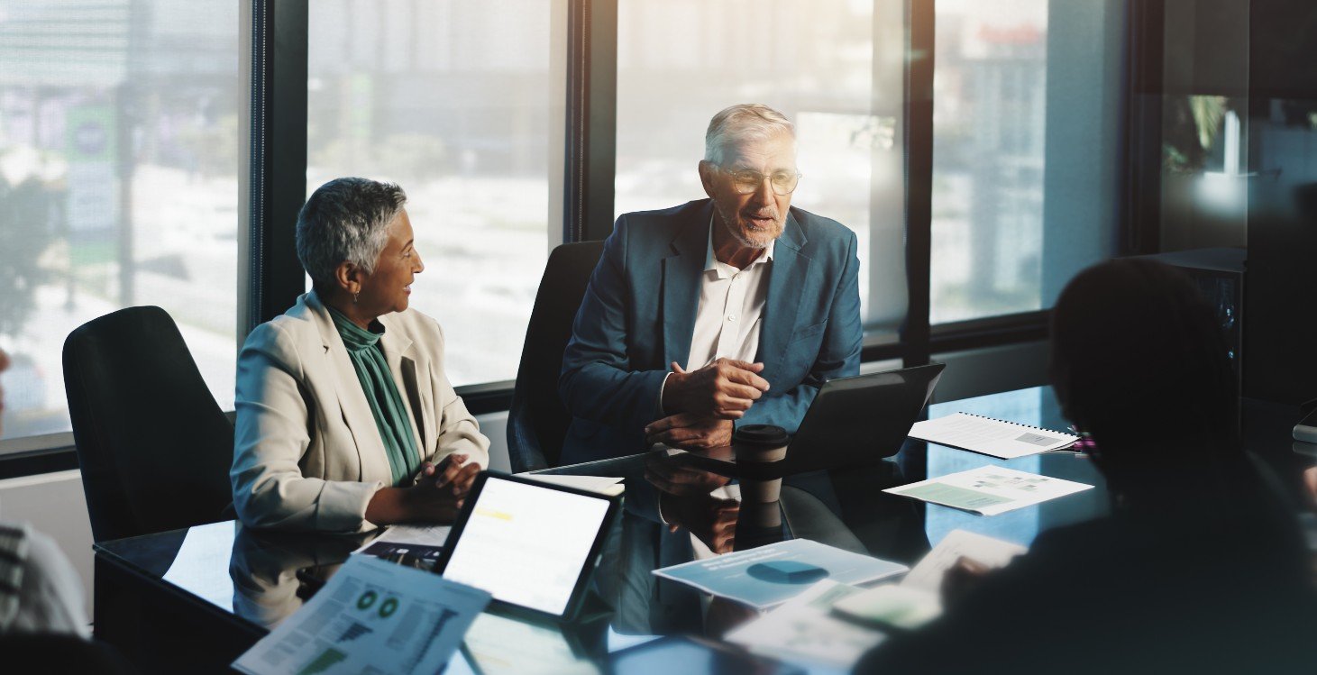 Two older business professionals in a meeting with other professionals sitting around a meeting table